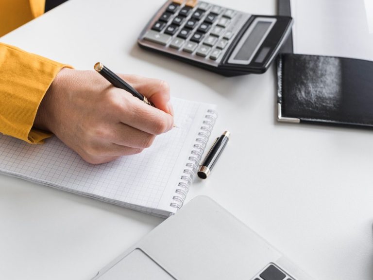 Main d'une personne écrivant dans un bloc-notes avec un stylo, calculatrice et documents.